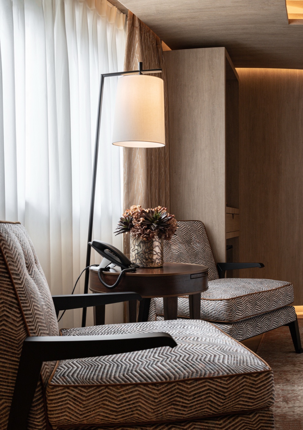 A warmly lit hotel lounge corner with patterned armchairs, a lamp and flowers on a side table