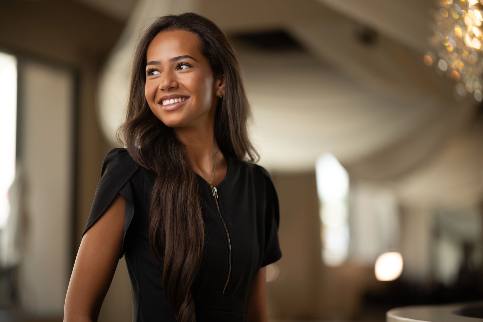 A smiling woman with long dark hair in a black dress, photographed in a bright venue with shallow depth of field