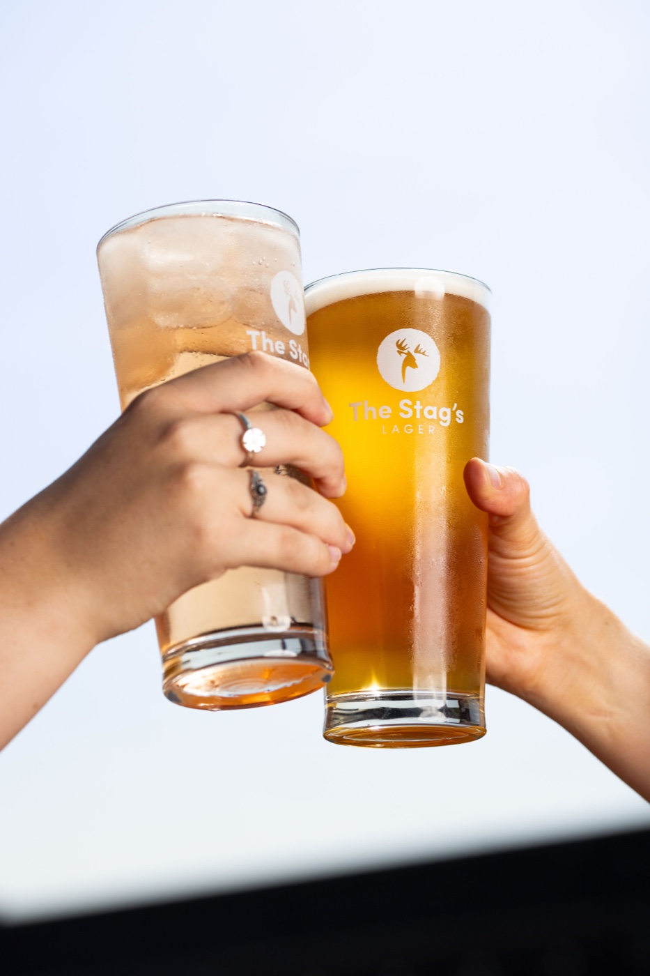 Two hands clinking pint glasses of lager and cider against a bright sky