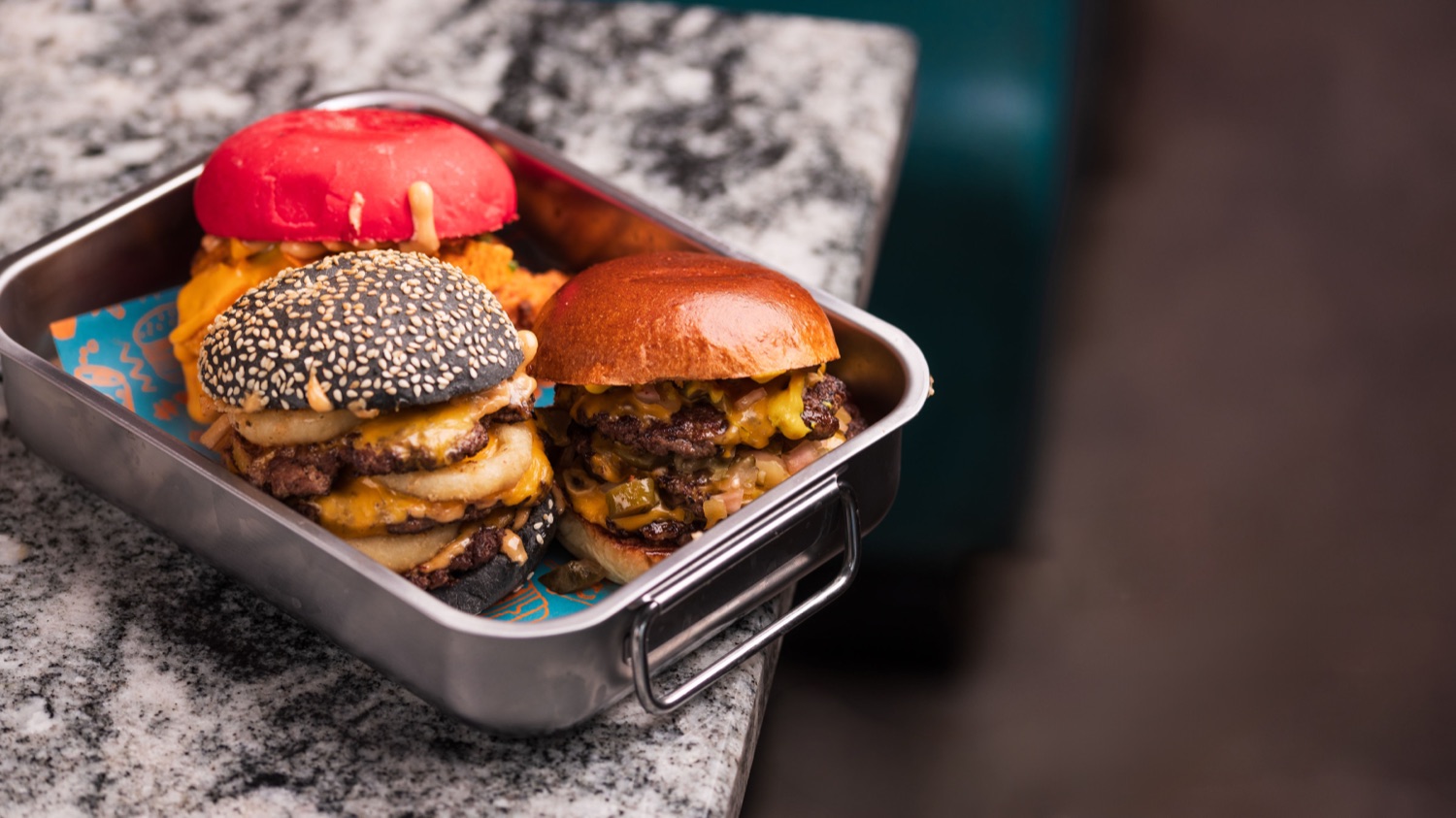 Three burgers in a steel serving tray at Burga Portswood, one with a red bun, one with a black sesame bun and one brioche, all stacked with cheese and onion rings, food photography by Ryan Stanikk