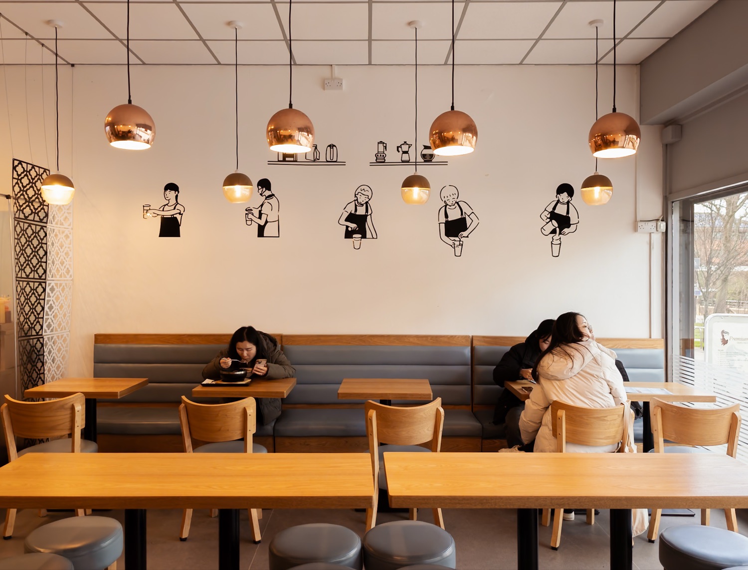 Interior of Mi Rice on Commercial Road Southampton with copper pendant lights, wall illustrations of people making drinks, pale wood tables and diners eating in the window
