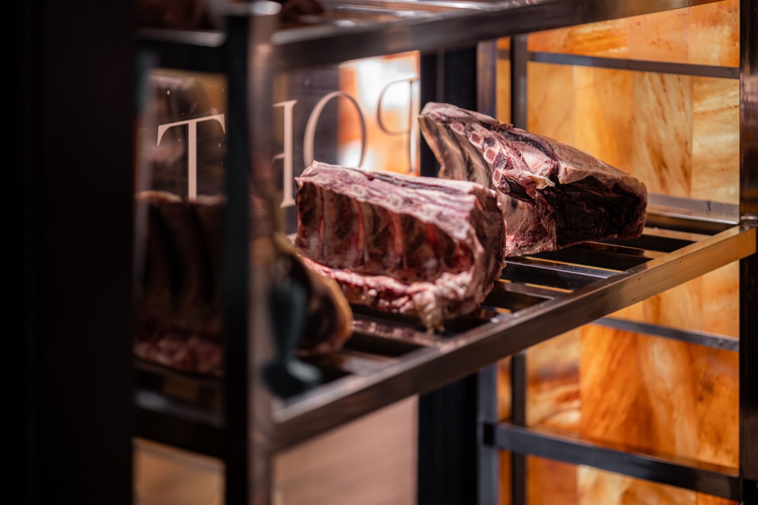 Bone-in beef rib joints dry ageing on steel racks inside the glass-fronted cabinet at Porters Steakhouse Southampton, lit by Himalayan salt panels, food photography by Ryan Stanikk
