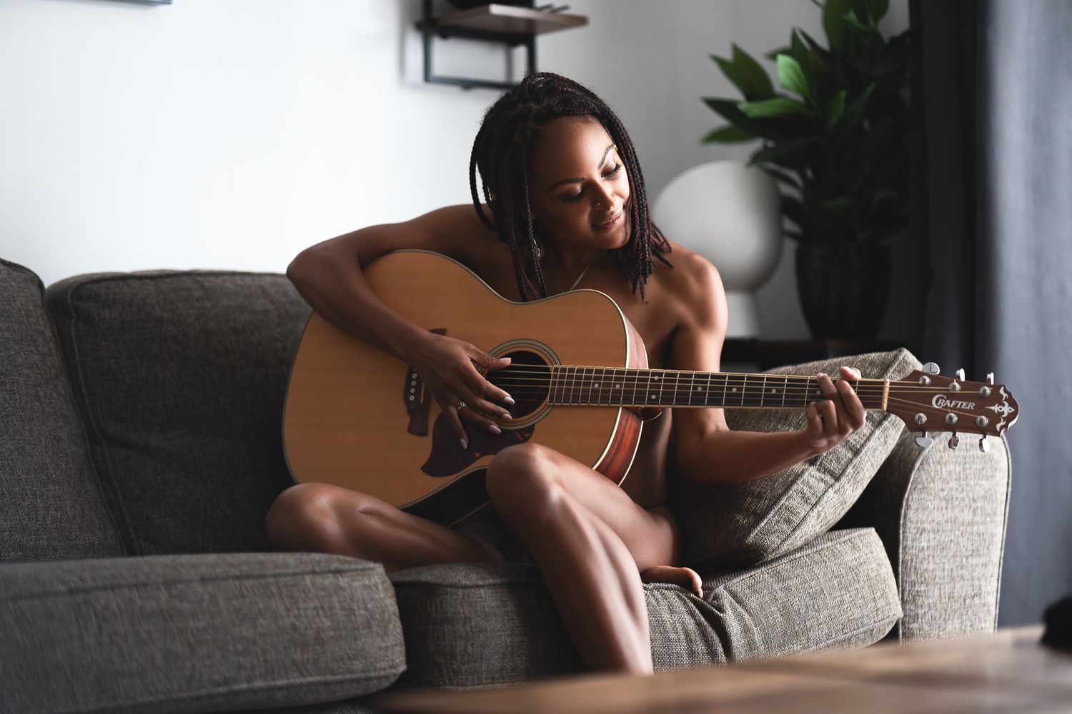 Woman with braids playing acoustic guitar on a grey sofa in soft natural light, relaxed creative boudoir photography Southampton