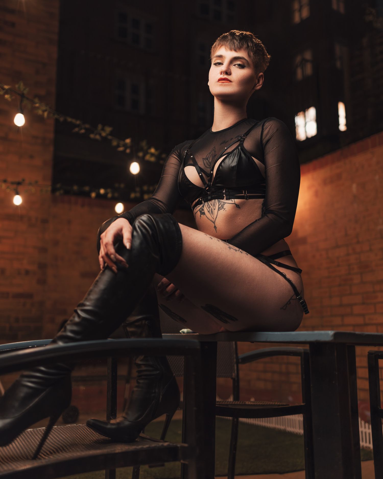 Short-haired woman in black mesh top and leather harness with knee-high boots seated on a rooftop railing at night with festoon lights, alternative urban boudoir photography Southampton