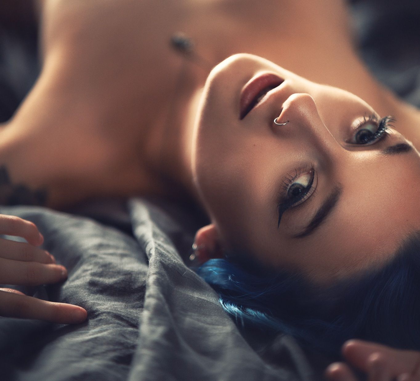 Close-up portrait of a woman with blue hair and nose piercing lying on grey bedding with warm lighting, intimate artistic boudoir photography Southampton