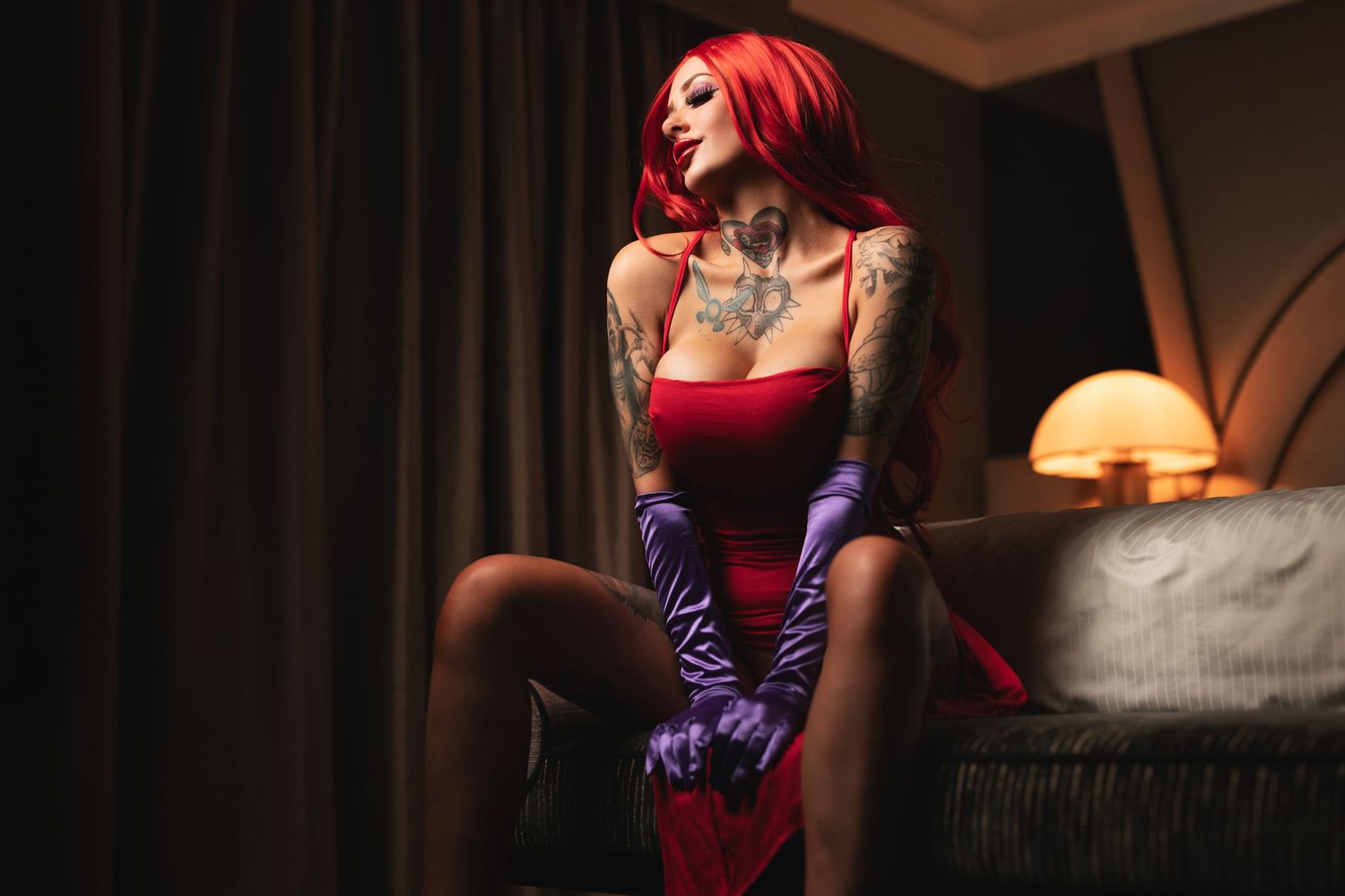 Tattooed woman with red hair in a red dress and purple satin gloves seated on a sofa with warm lamplight, creative themed boudoir photography Southampton