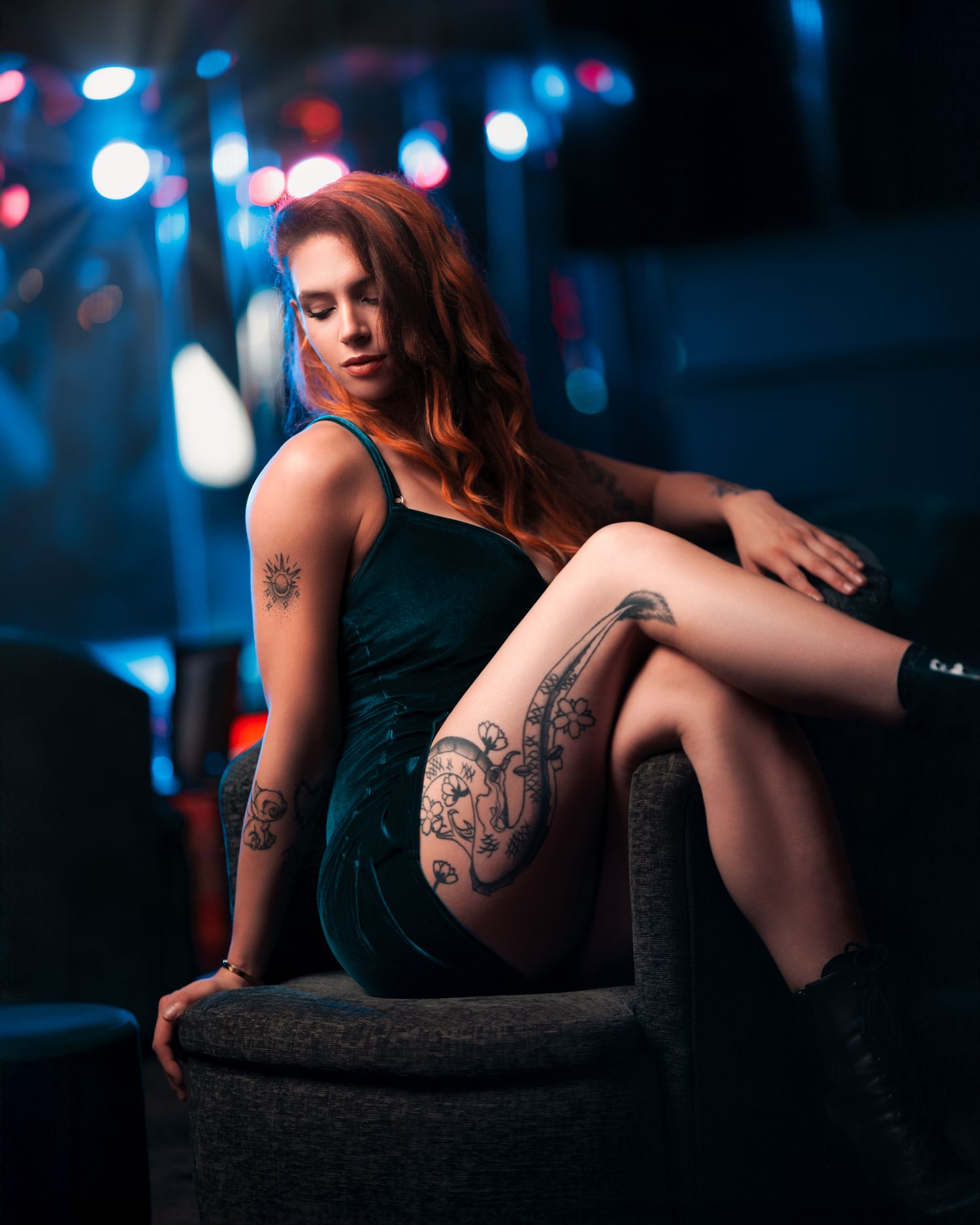 Tattooed woman with auburn hair in a dark green velvet dress seated in a moody setting with colourful bokeh lights, creative styled boudoir photography Southampton