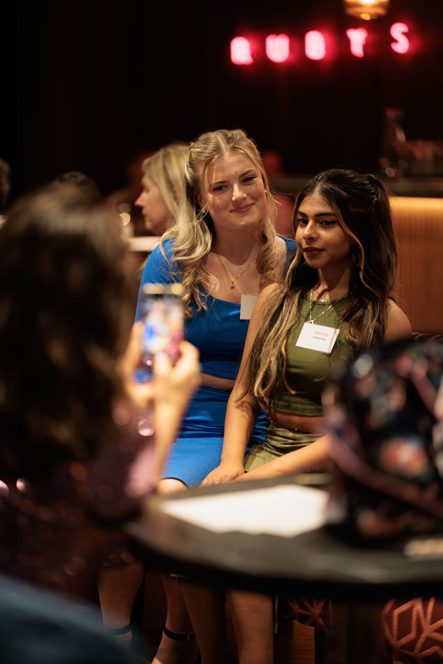 Two young women with name badges posing for a photo at a corporate networking event at a bar with neon signage, corporate event photography