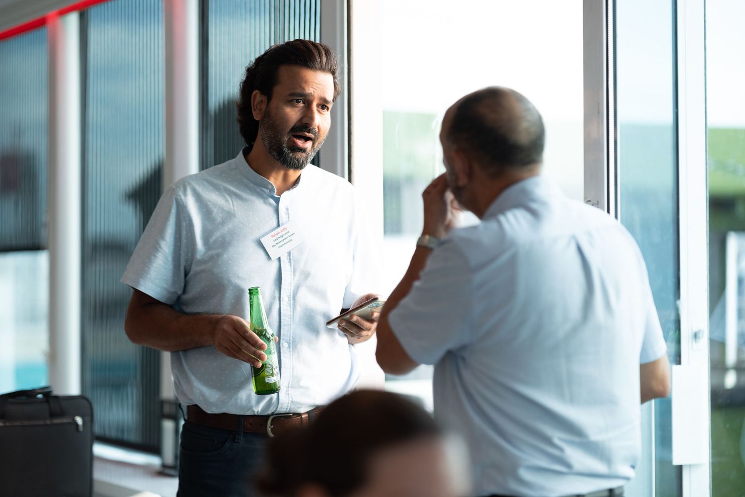 Two men in conversation with drinks at a corporate networking event by window light, business event photography