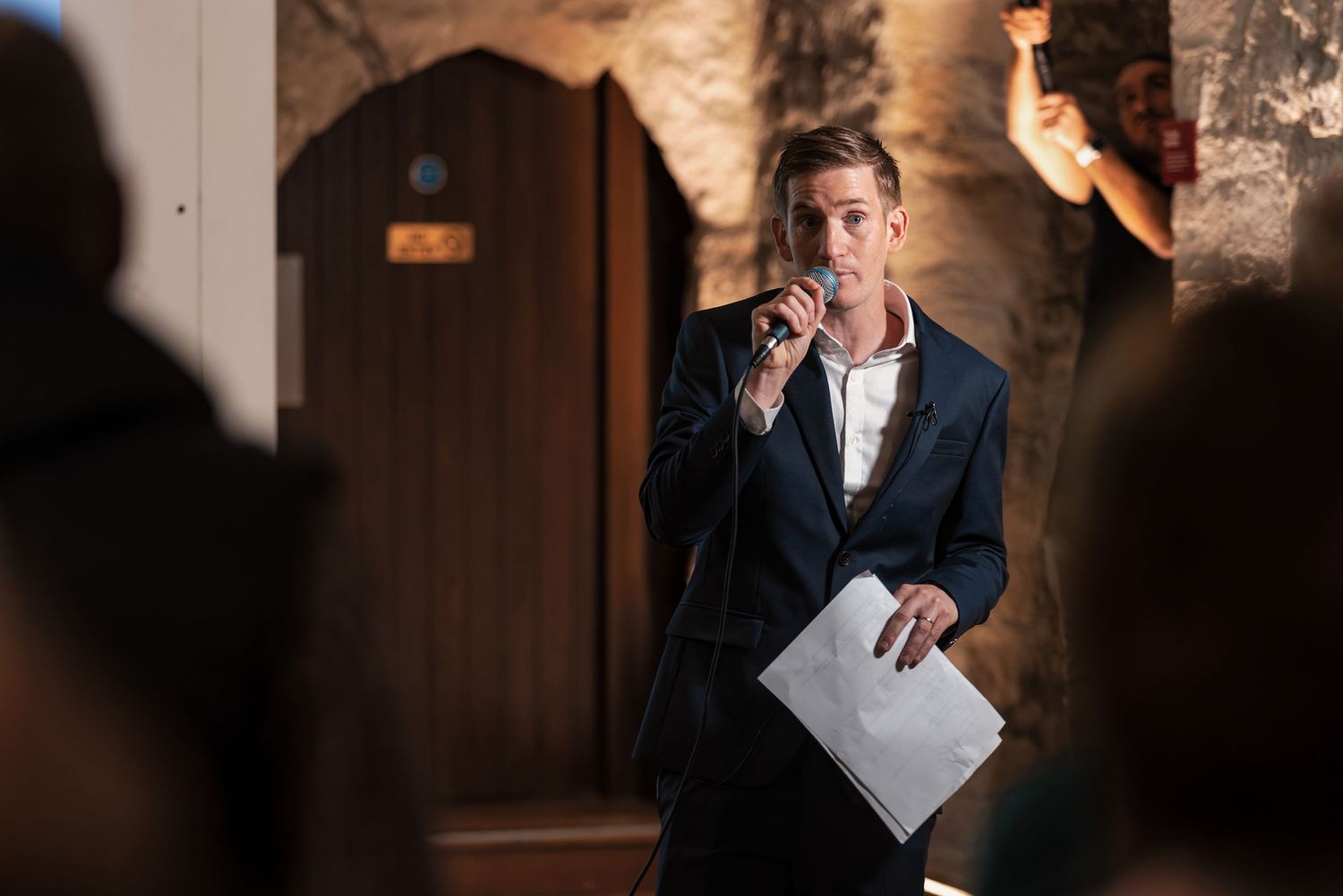 Host in a suit speaking into a microphone at a corporate event in a stone-walled venue, corporate event speaker photography