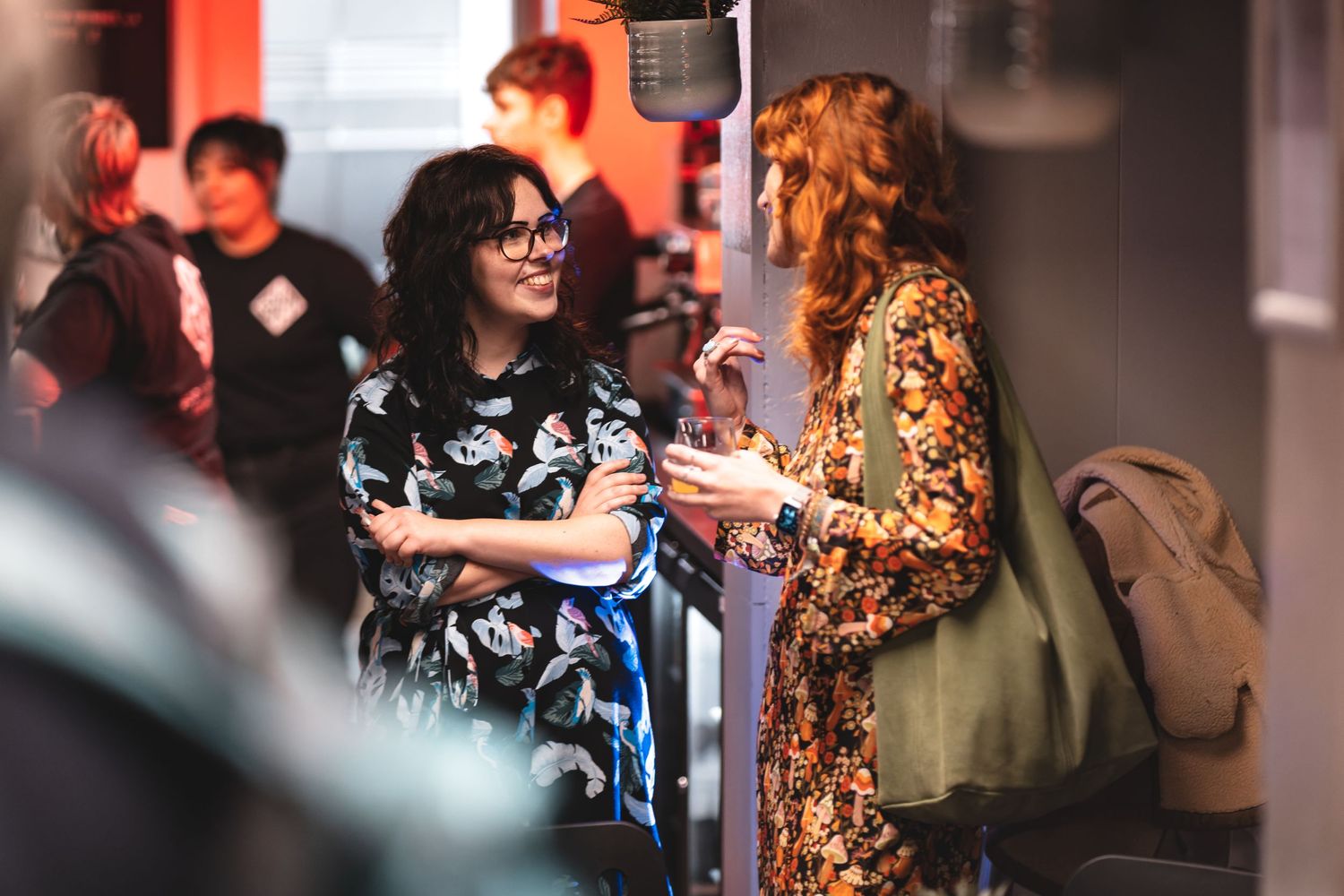 Two women chatting with drinks at a networking event with warm ambient lighting, candid corporate event photography