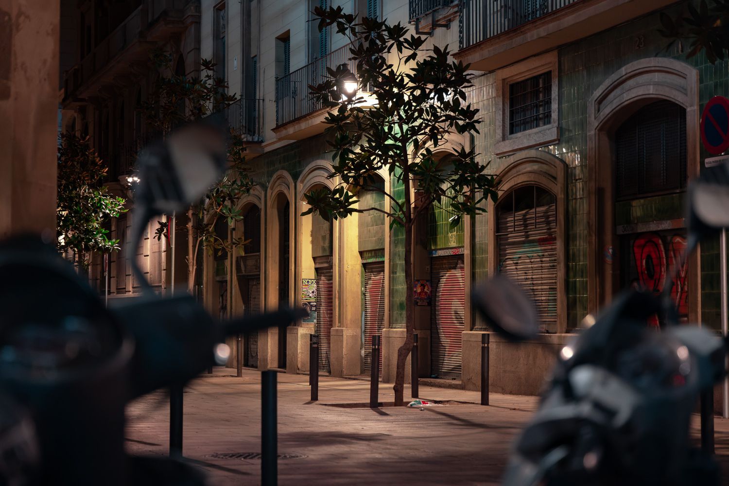 European city street at night with ornate stone building facades, street lights, trees, parked motorcycles in foreground and lone pedestrian, architectural exterior photography