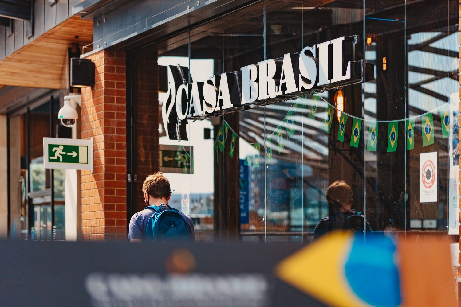 Casa Brasil restaurant exterior with large glass windows, brick facade, Brazilian flag bunting and pedestrians passing, commercial exterior photography