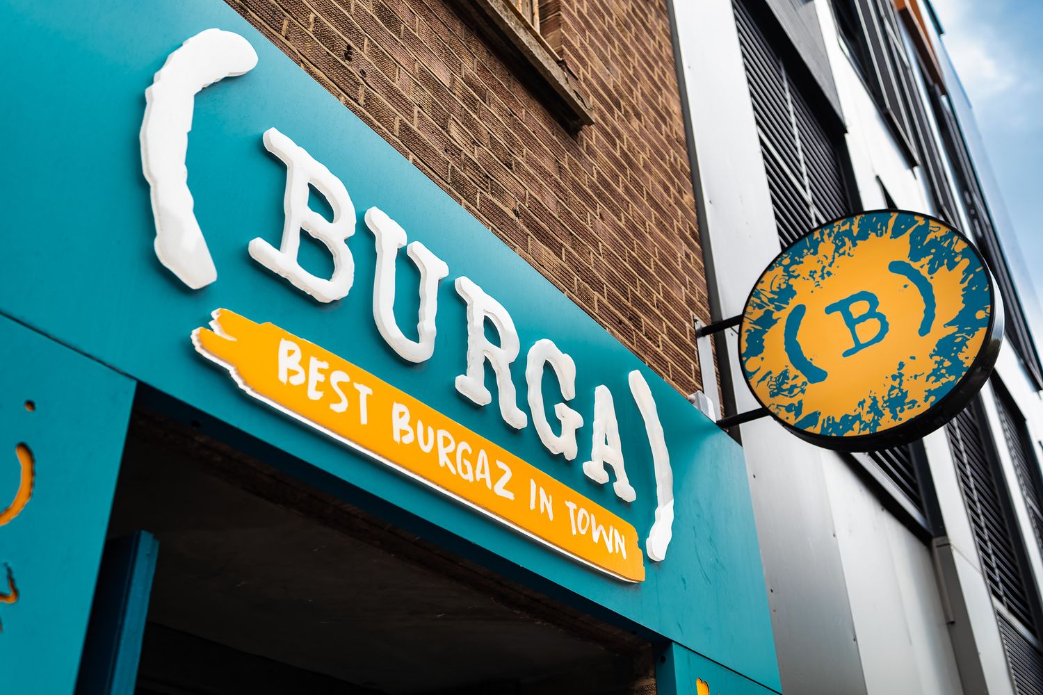 Burga restaurant teal facade with white 3D lettering, Best Burgaz In Town tagline, and circular orange hanging sign against brick building exterior, commercial signage photography