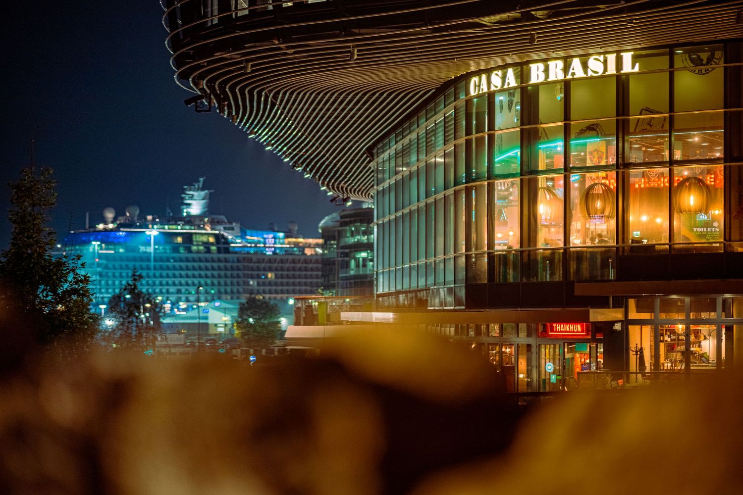 Casa Brasil restaurant exterior at night with illuminated signage, curved glass facade, warm interior glow and cruise ship in the background, commercial dusk photography