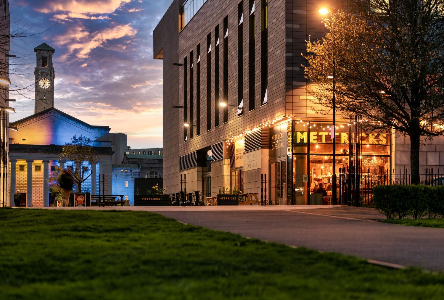 Mettricks Guildhall cafe exterior at twilight with illuminated signage and festoon lights, Southampton Guildhall clock tower, green lawn and dramatic sunset sky, commercial dusk photography
