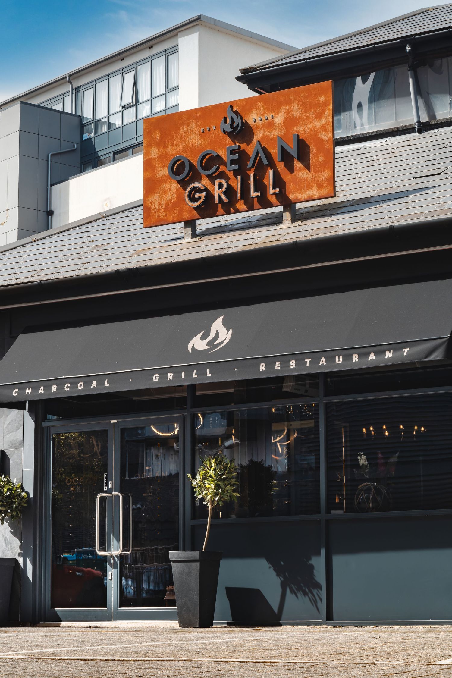 Ocean Grill restaurant exterior with large orange sign, dark grey awning reading Charcoal Grill Restaurant, glass windows and potted plant, commercial exterior photography