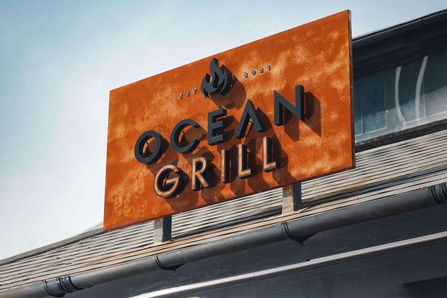 Ocean Grill orange corten steel sign with flame logo mounted on building roofline against clear sky, close-up commercial signage photography