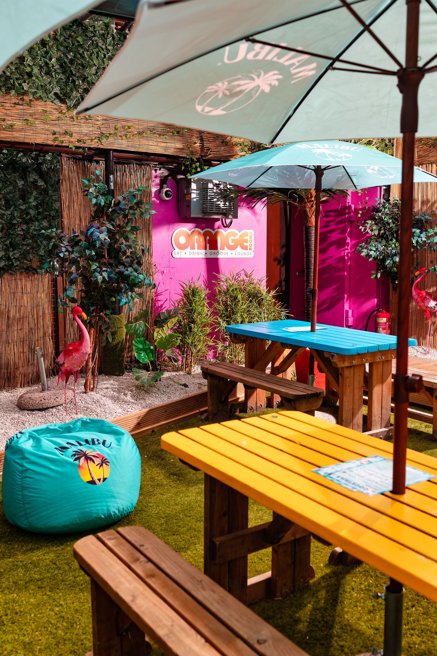 Orange Rooms colourful outdoor bar area with parasols, yellow and wooden picnic benches, pink container with Orange signage, teal Malibu beanbag and artificial grass, commercial exterior photography