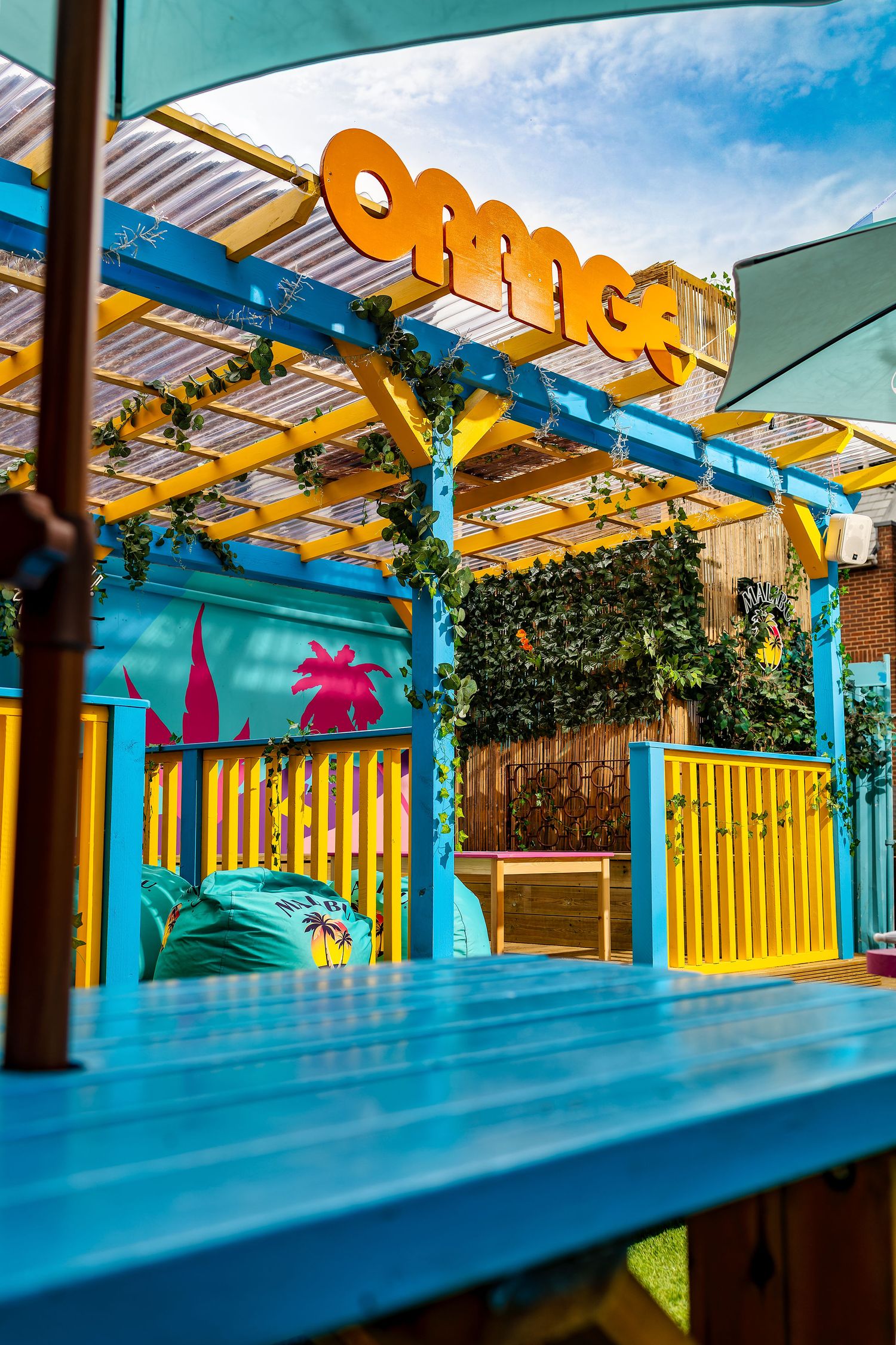 Orange Rooms outdoor area with orange 3D lettering, yellow and blue pergola, climbing plants, teal table and Malibu beanbag, colourful exterior photography