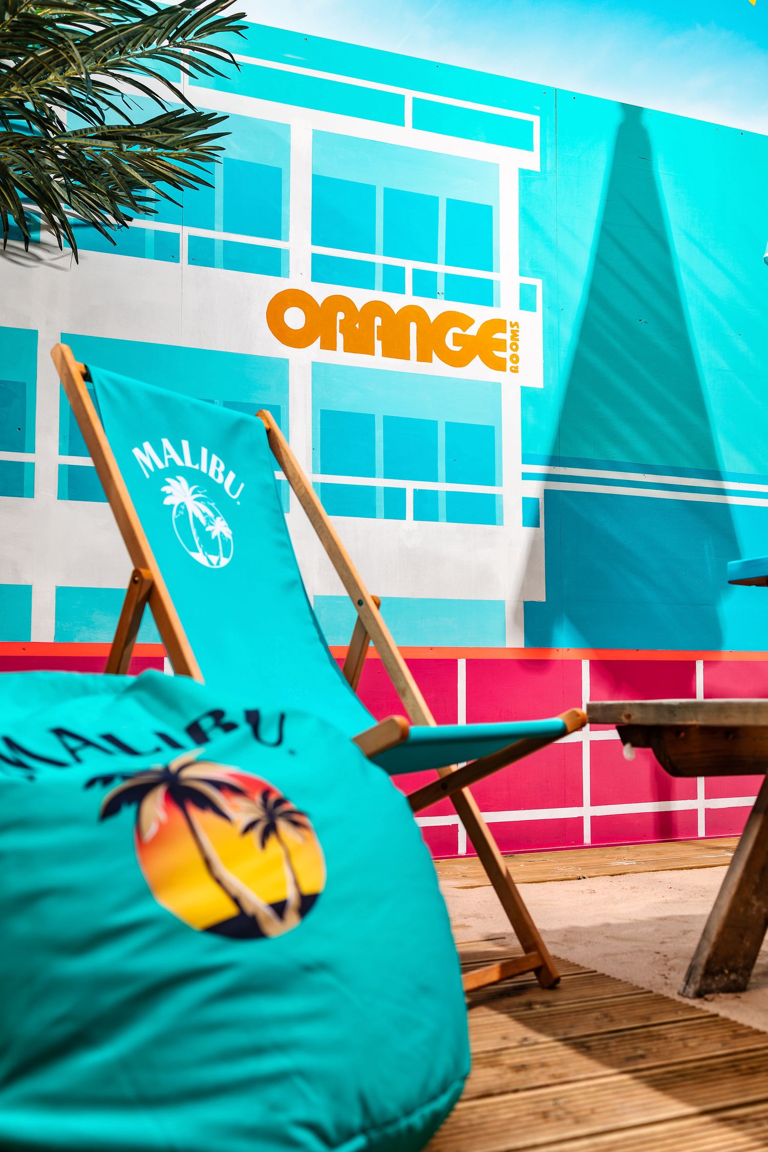 Orange Rooms outdoor area with teal wall mural, orange signage, Malibu-branded beanbag and deckchair on wooden decking, colourful exterior photography