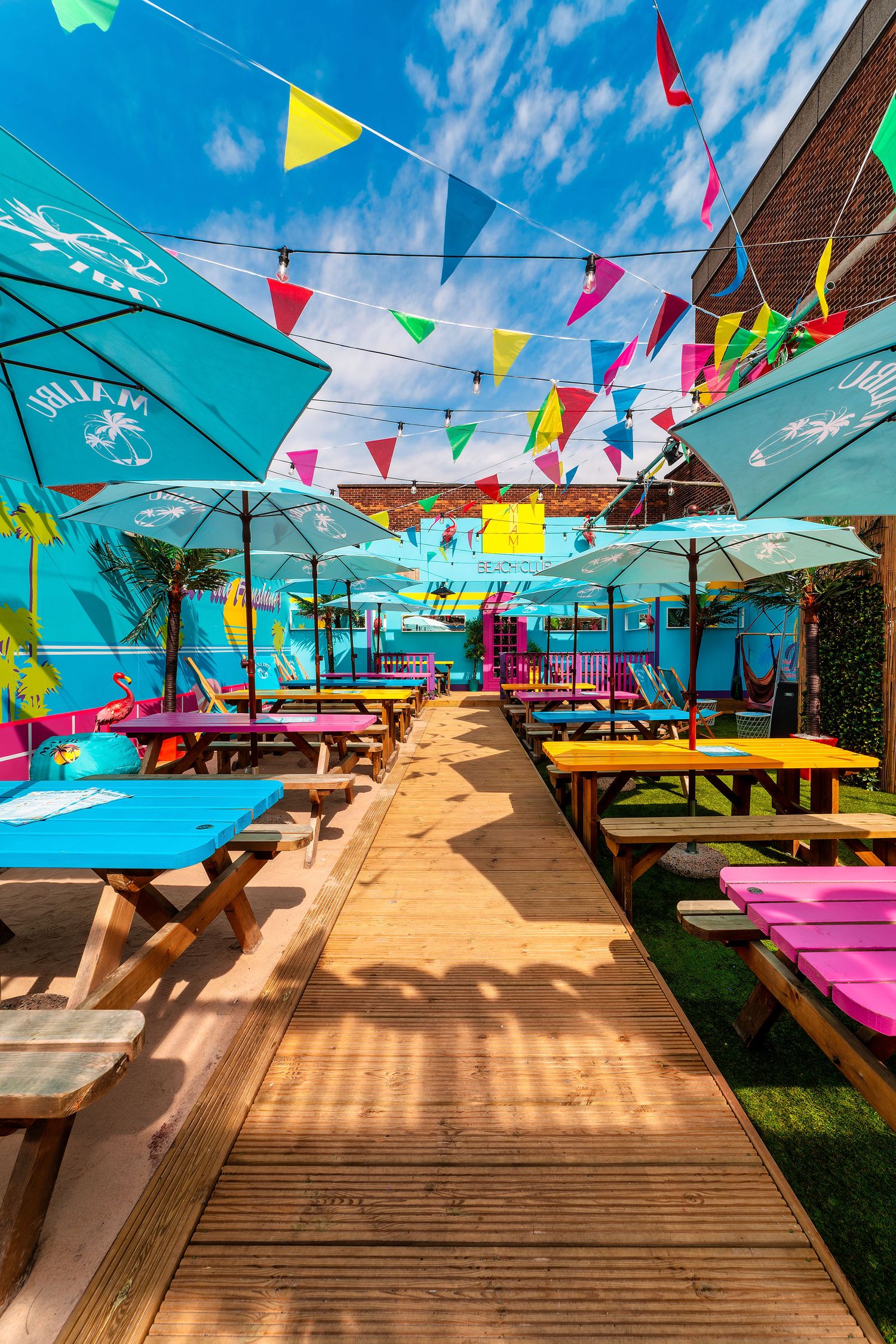 Orange Rooms colourful outdoor beer garden with teal parasols, bright bunting, painted picnic benches in blue, yellow and pink, and wooden decking walkway, commercial exterior photography