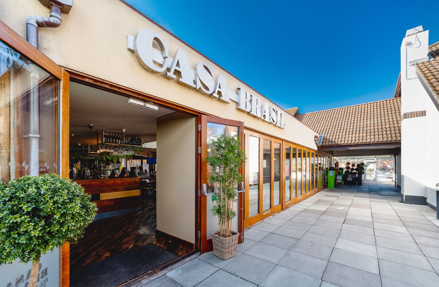 Casa Brasil Portsmouth exterior with rooftop signage, open wooden bi-fold doors, potted plants and paved walkway under blue sky, commercial exterior photography