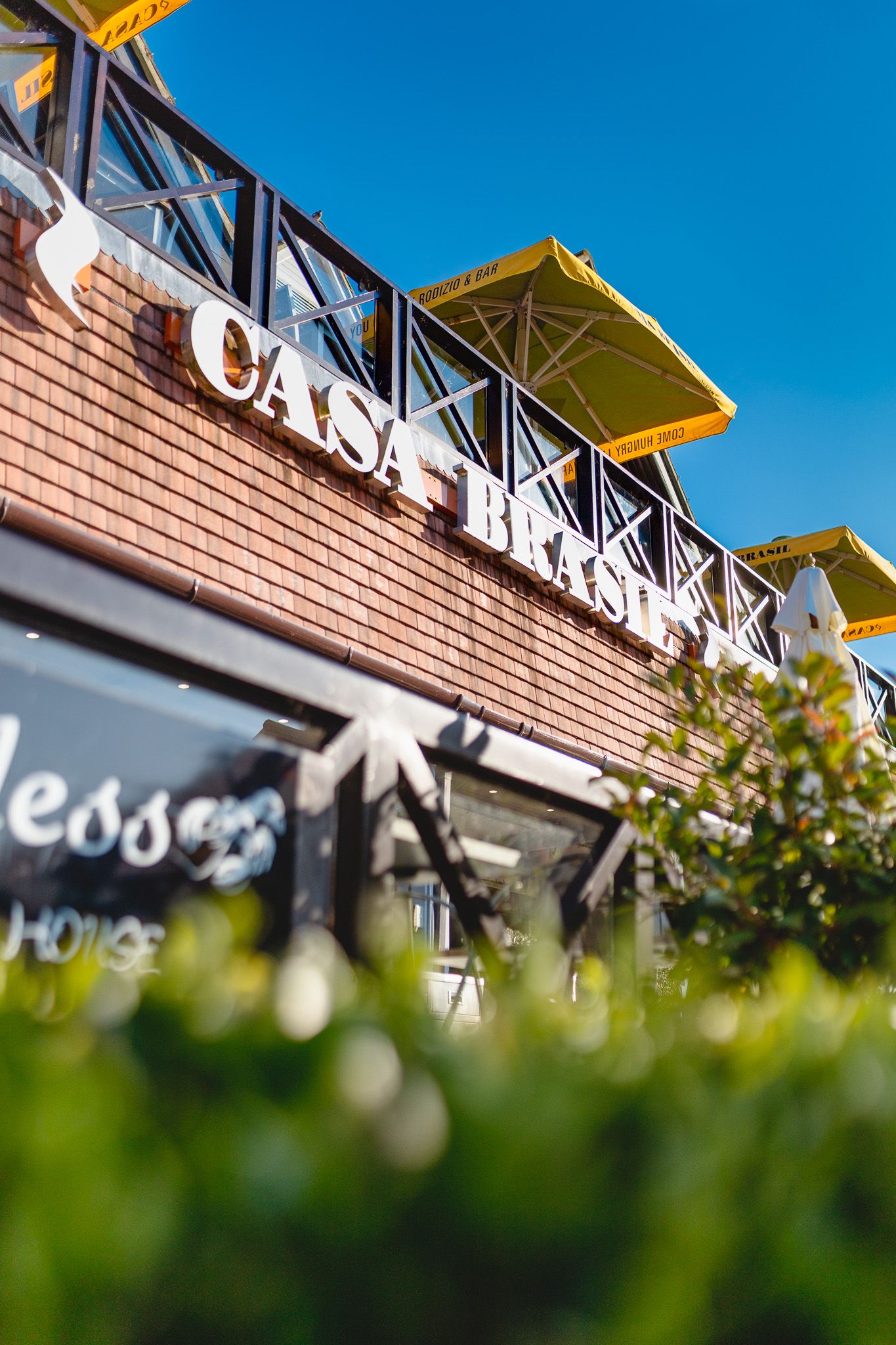 Casa Brasil building exterior with yellow parasols on upper level, brick facade and rooftop signage against blue sky with foreground greenery, architectural exterior photography