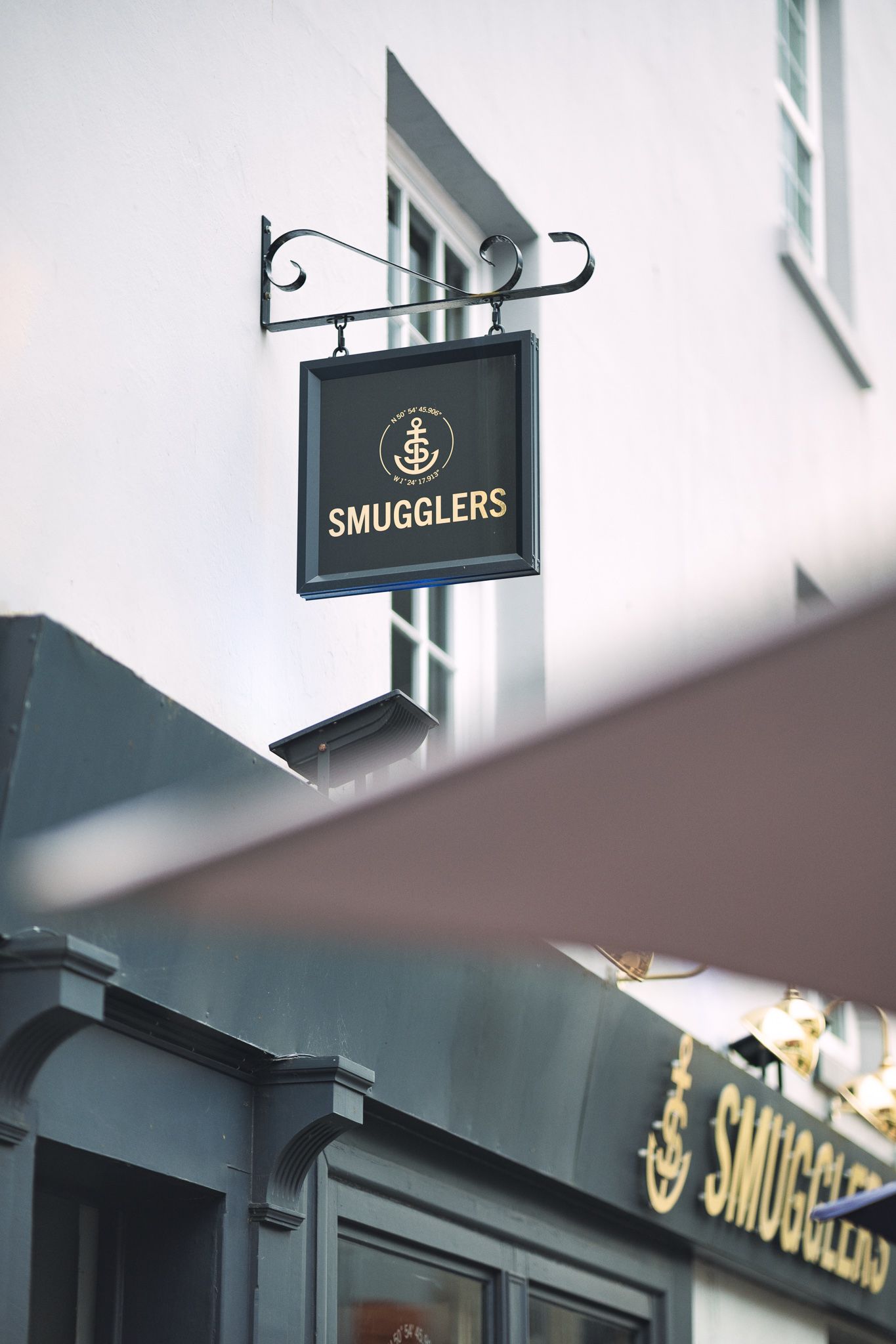 Smugglers bar hanging sign with anchor logo on wrought iron bracket against white building exterior, with dark shopfront below, commercial signage photography