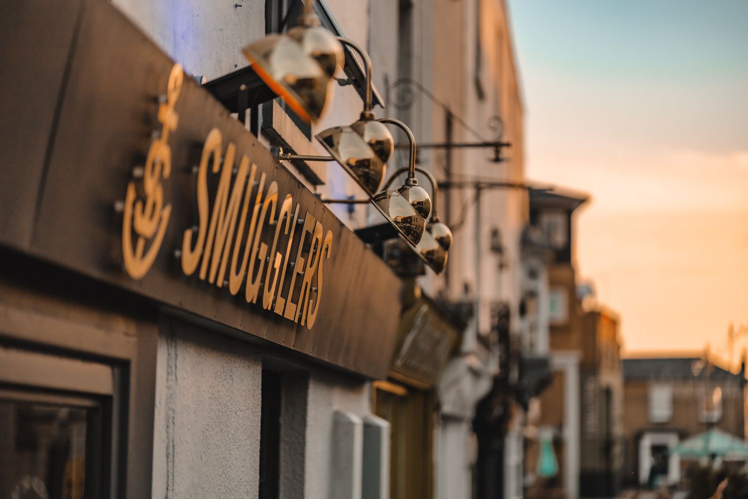 Smugglers bar exterior signage close-up at golden hour with brass gooseneck lights, gold lettering and anchor logo against warm sunset sky, commercial twilight photography