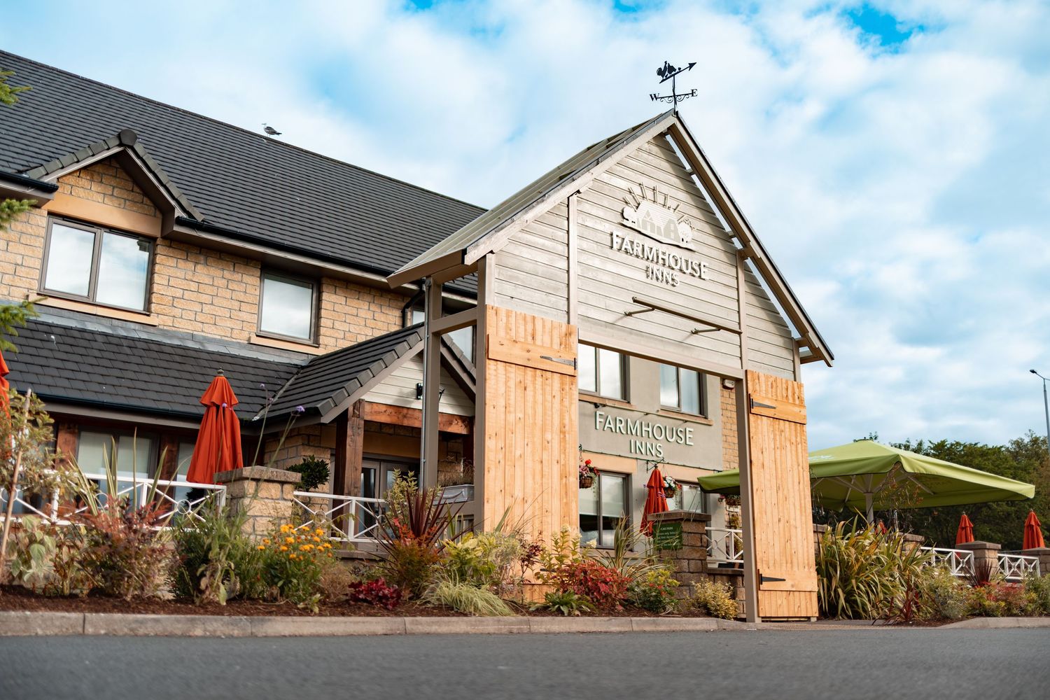 Farmhouse Inns pub exterior with timber cladding, grey roof, weather vane, red parasols and landscaped gardens under blue sky, commercial exterior photography