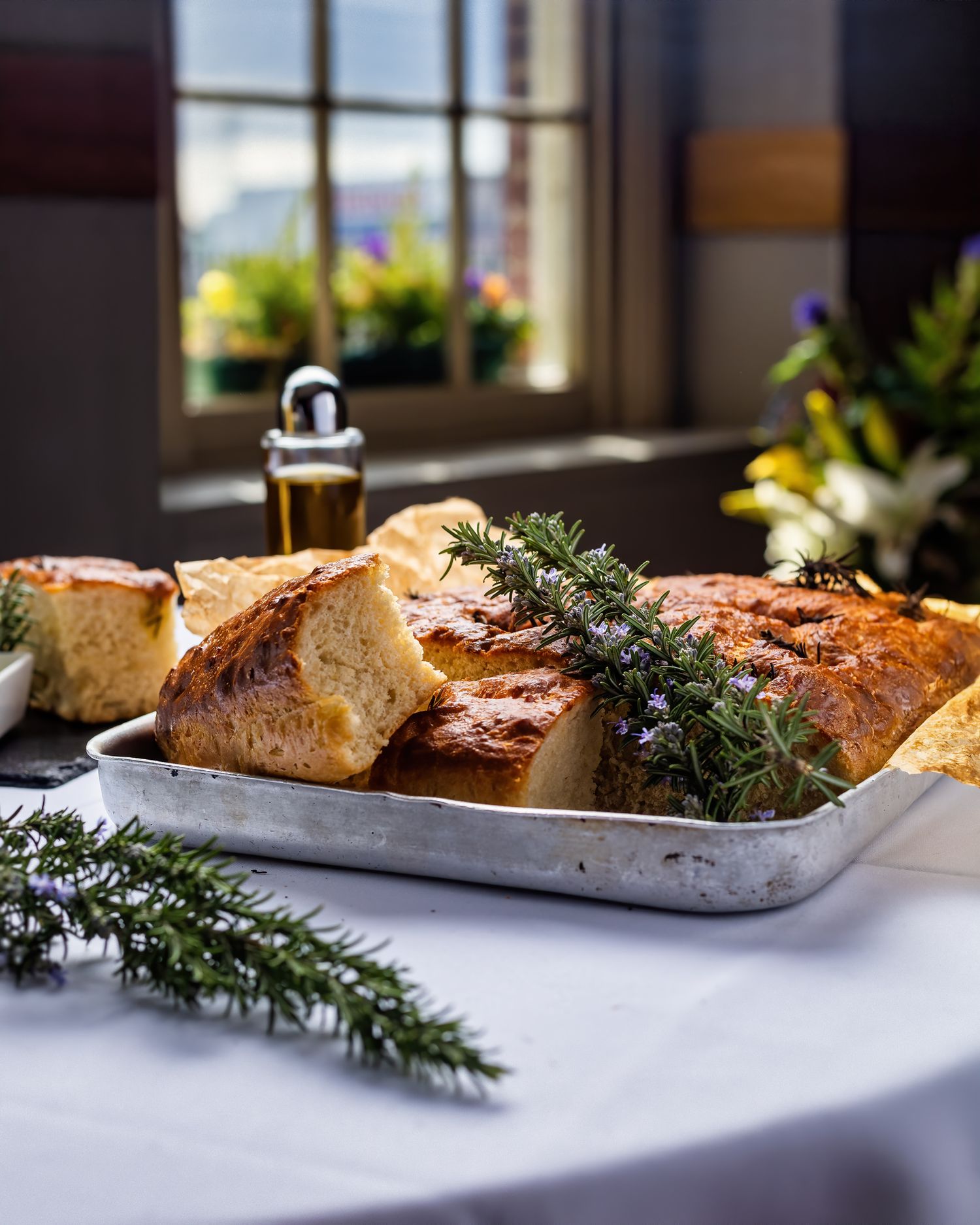 Rustic focaccia bread with fresh rosemary sprigs on a metal tray with olive oil and natural window light at Ennios Italian restaurant, food photography