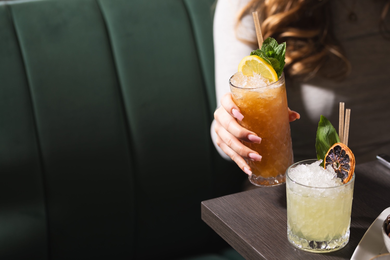 Hand holding a crushed ice cocktail with lemon and mint garnish beside a second cocktail with dried orange at Figurati Italian restaurant Southampton, drink photography