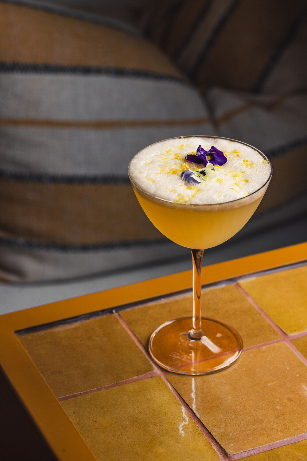 Pale yellow cocktail with an edible flower garnish on a mustard tiled table at Ixchel Mexican restaurant, King's Road Chelsea, London drink photography