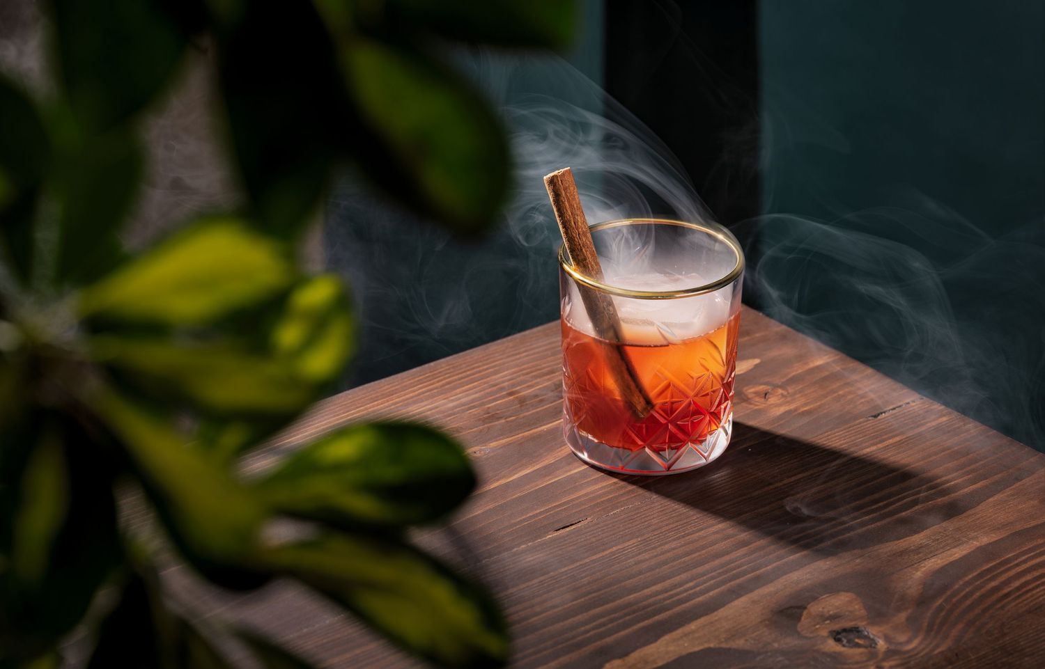 Smoky cocktail with a cinnamon stick garnish in a crystal cut rocks glass on a wooden bar with greenery at Kesarum restaurant Southampton, cocktail photography