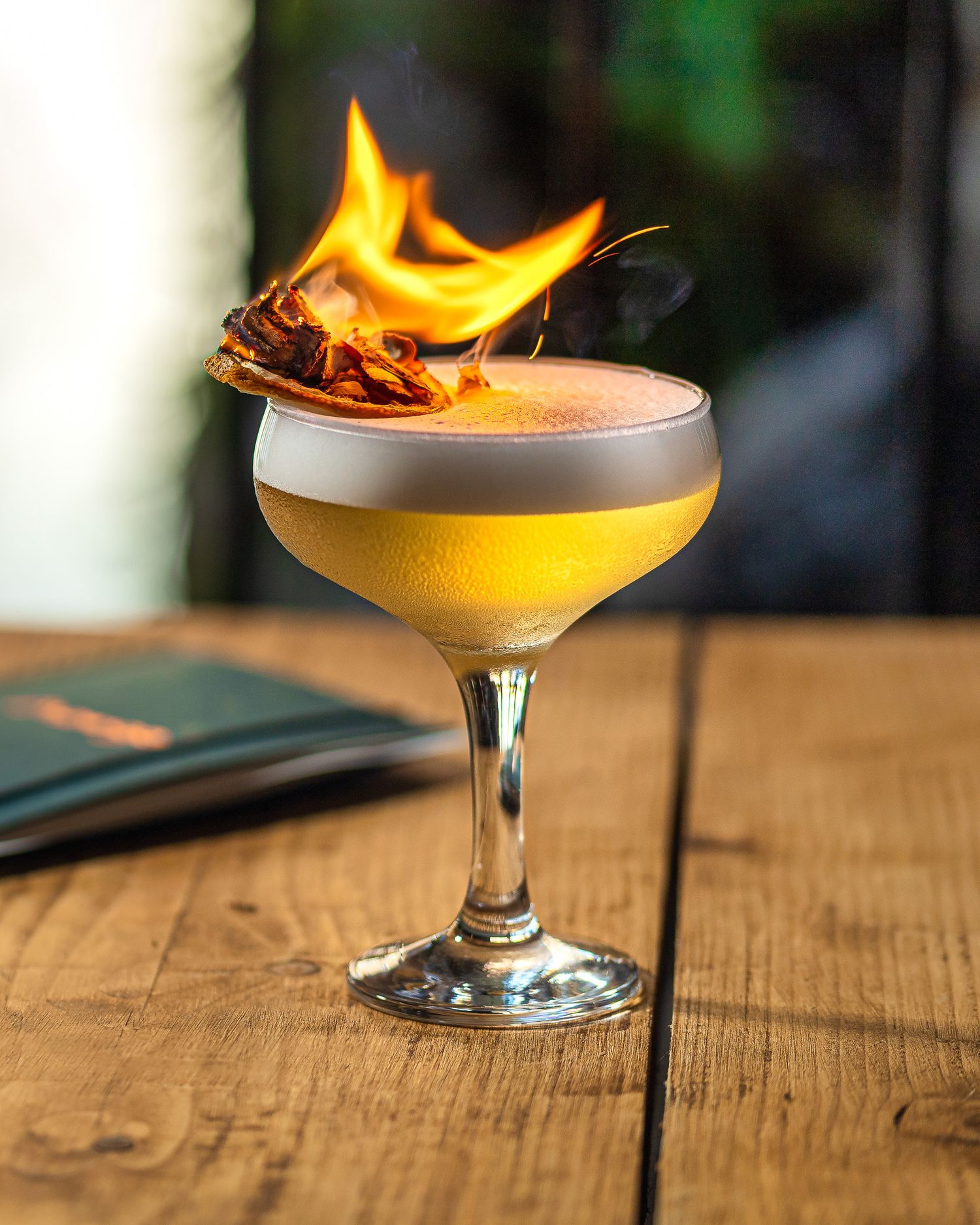 Flaming golden sour cocktail with egg white foam and a torched orange garnish in a coupe glass on a wooden table at Mango restaurant Southampton, cocktail photography