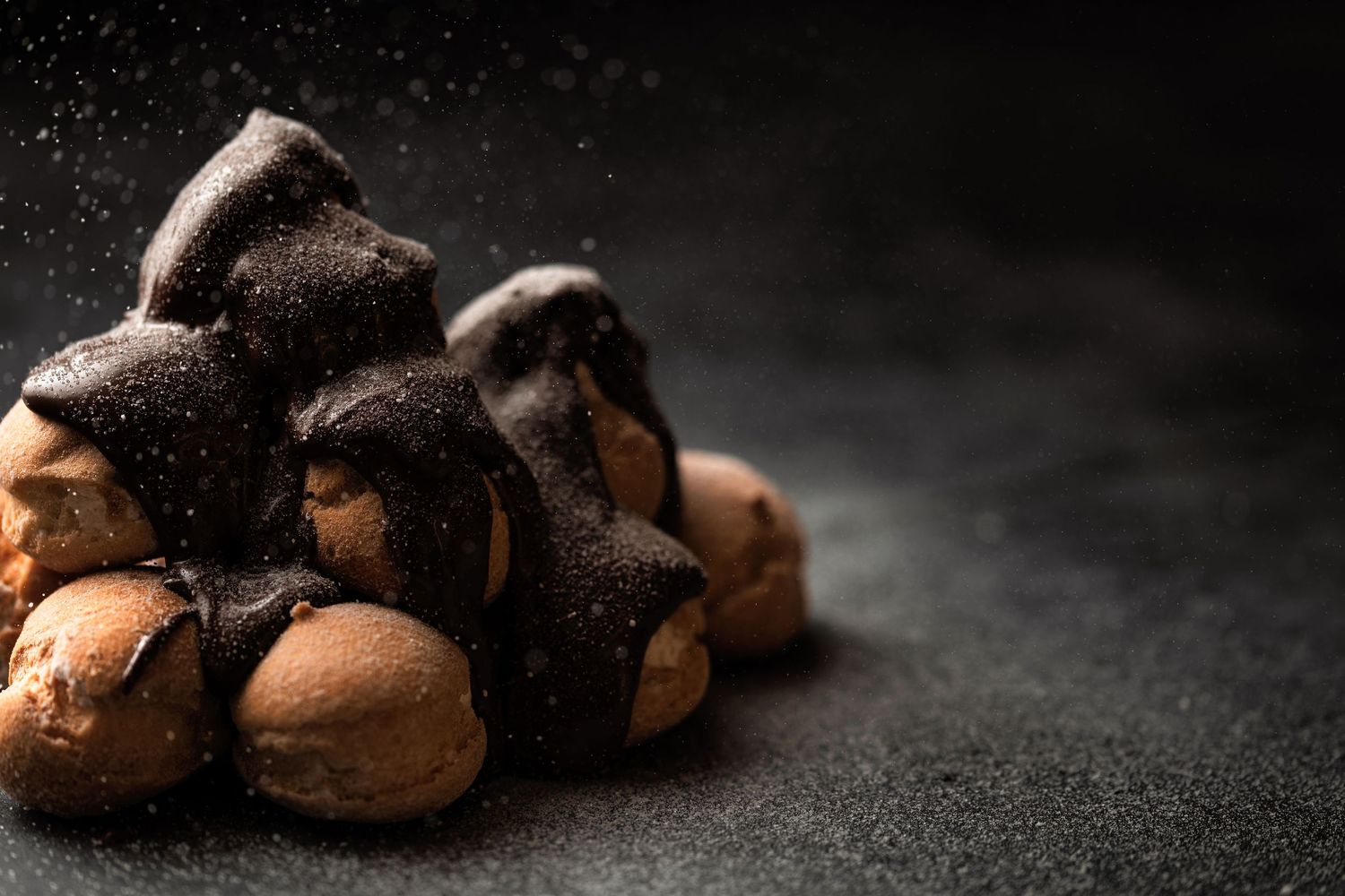 Chocolate profiteroles drizzled with dark chocolate sauce and dusted with icing sugar on a dark moody background, artisan dessert patisserie food photography