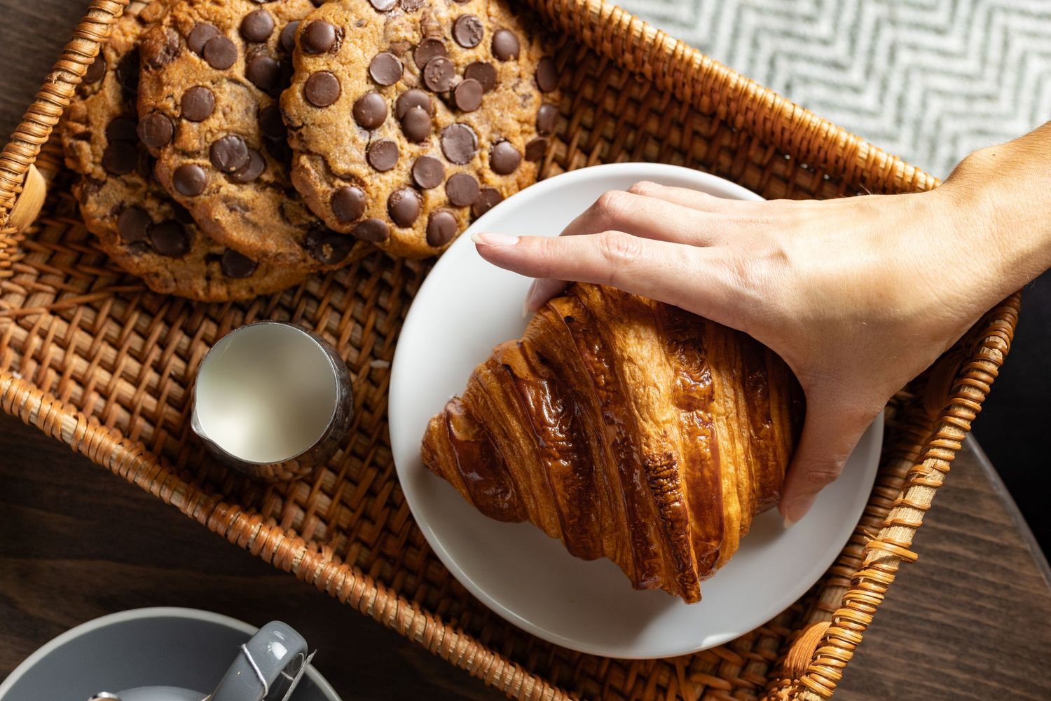 Hand reaching for a golden croissant alongside chocolate chip cookies and milk in a woven basket tray at Room2 cafe Southampton, bakery food photography