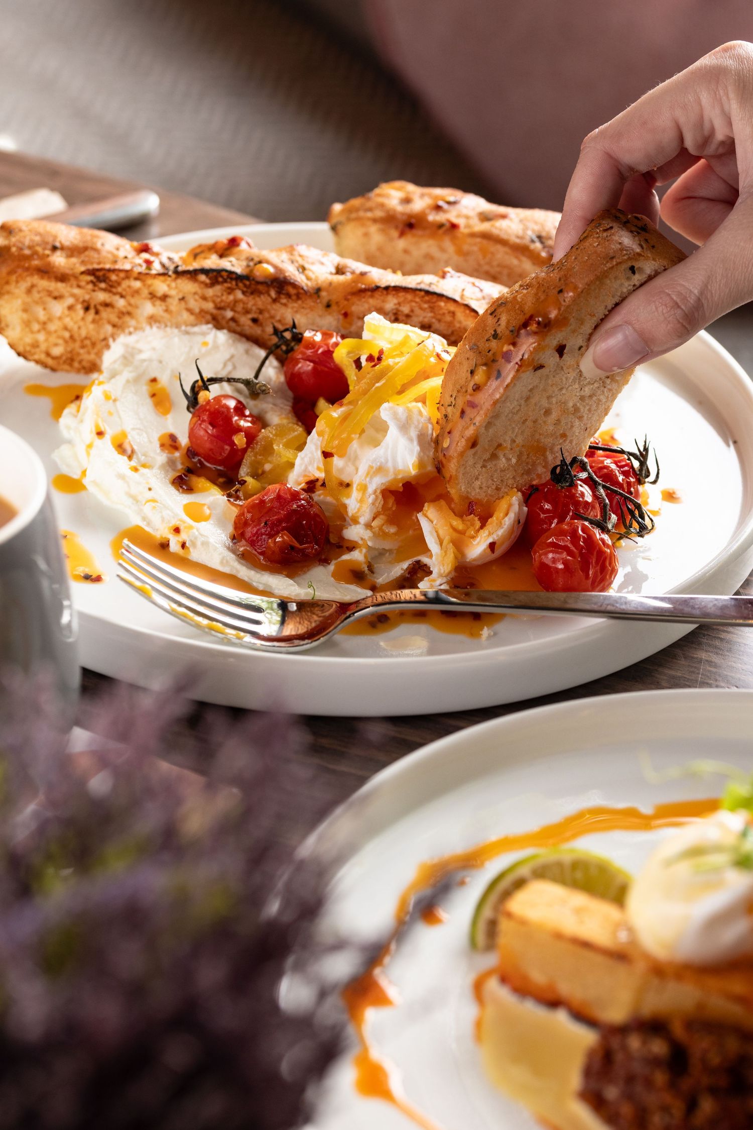 Hand dipping toast into poached eggs with roasted cherry tomatoes and chilli oil in natural light, brunch food photography