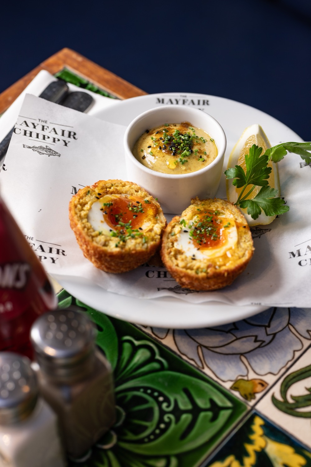 Scotch eggs halved to show soft-boiled centre with curry mayo dipping sauce, lemon wedge and fresh parsley on branded Mayfair Chippy greaseproof paper, food photography London