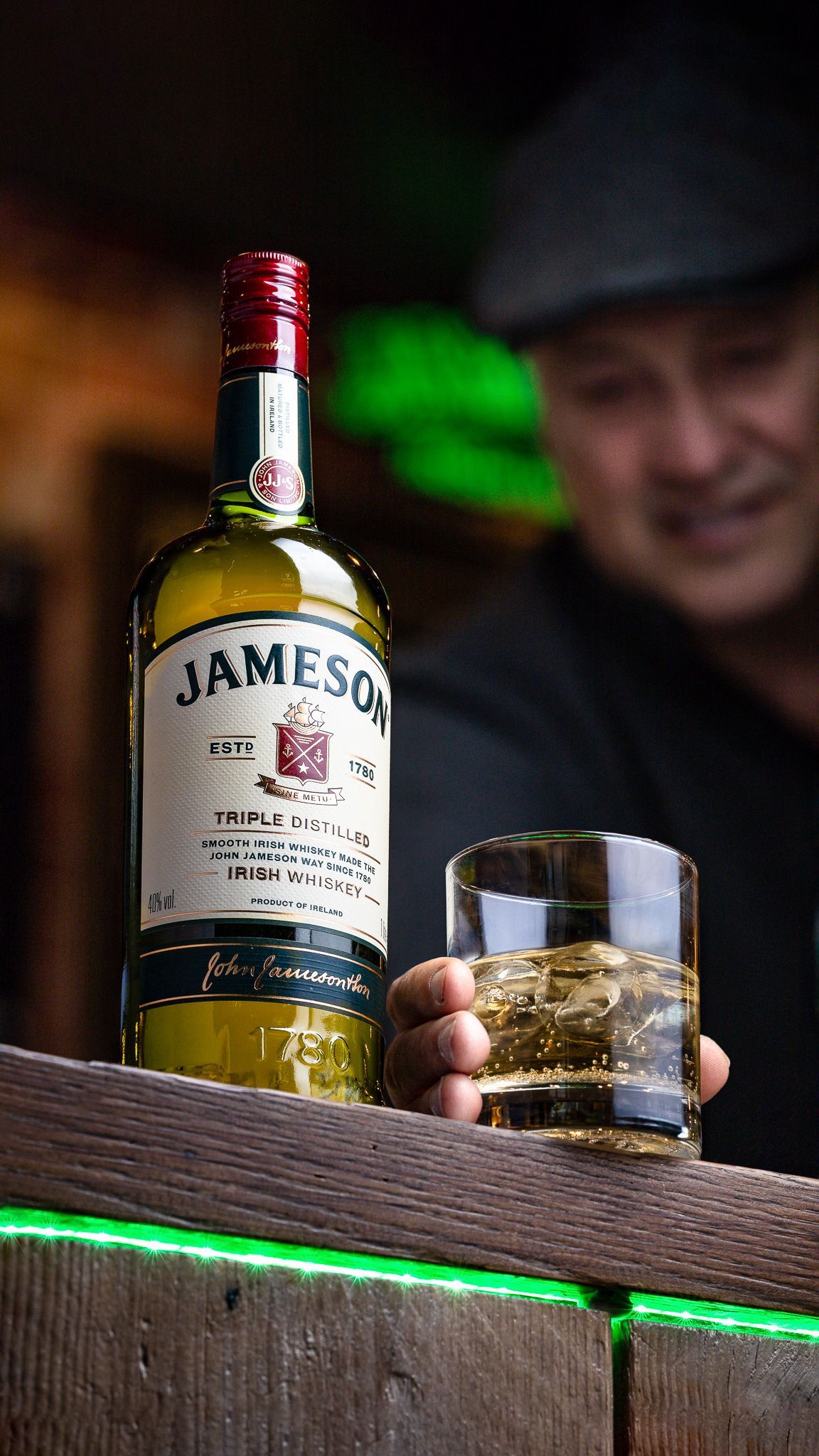 Jameson Irish whiskey bottle with a man holding a glass on the rocks at Shenanigans pub with green neon lighting, whiskey drink photography Southampton