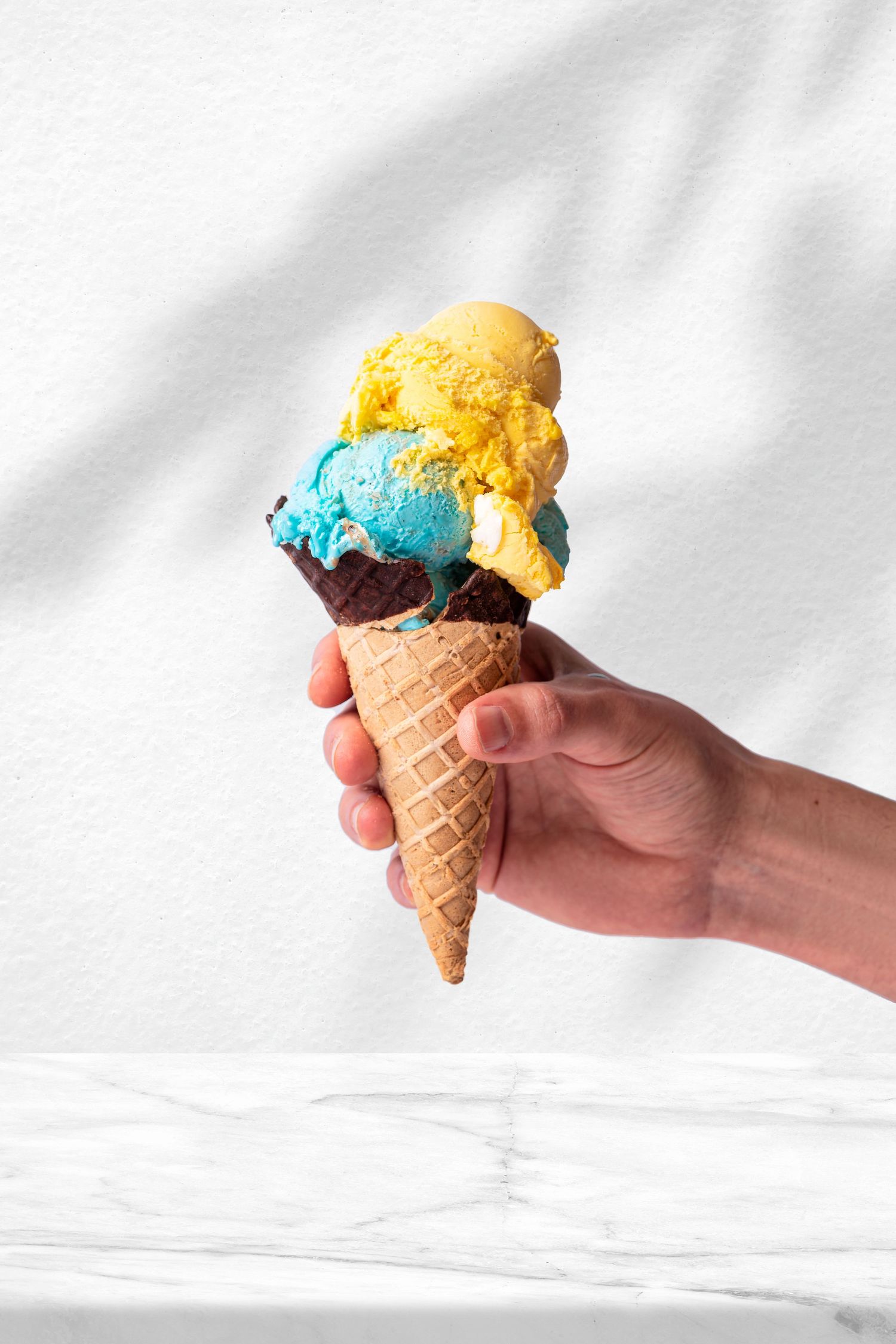 Hand holding colourful gelato scoops in blue, yellow and chocolate in a waffle cone against a white wall at Sprinkles ice cream shop, dessert photography