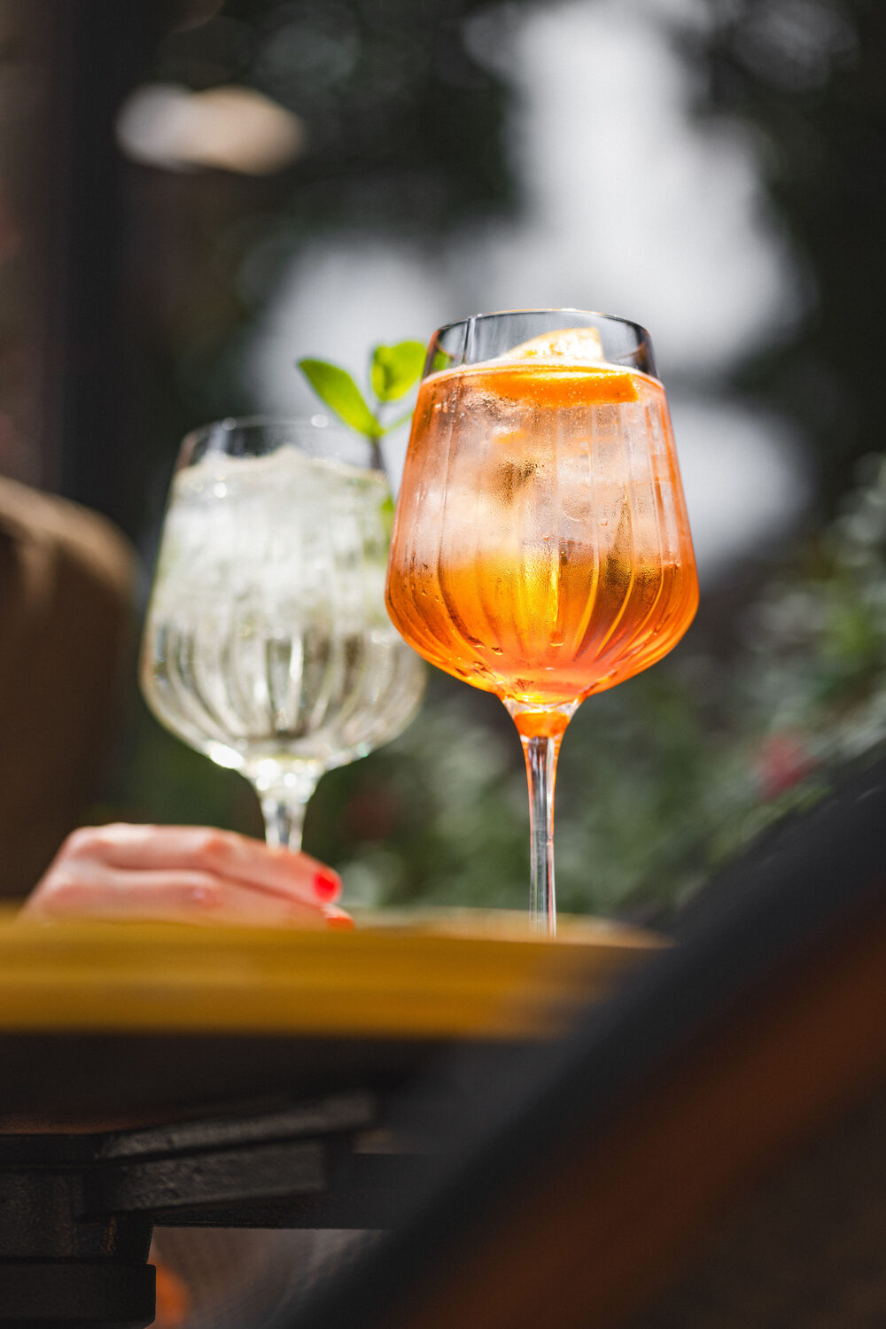 Aperol spritz in a ribbed stemmed glass with a second drink behind on a sunlit terrace table at The Trafalgar Chelsea, London drink photography