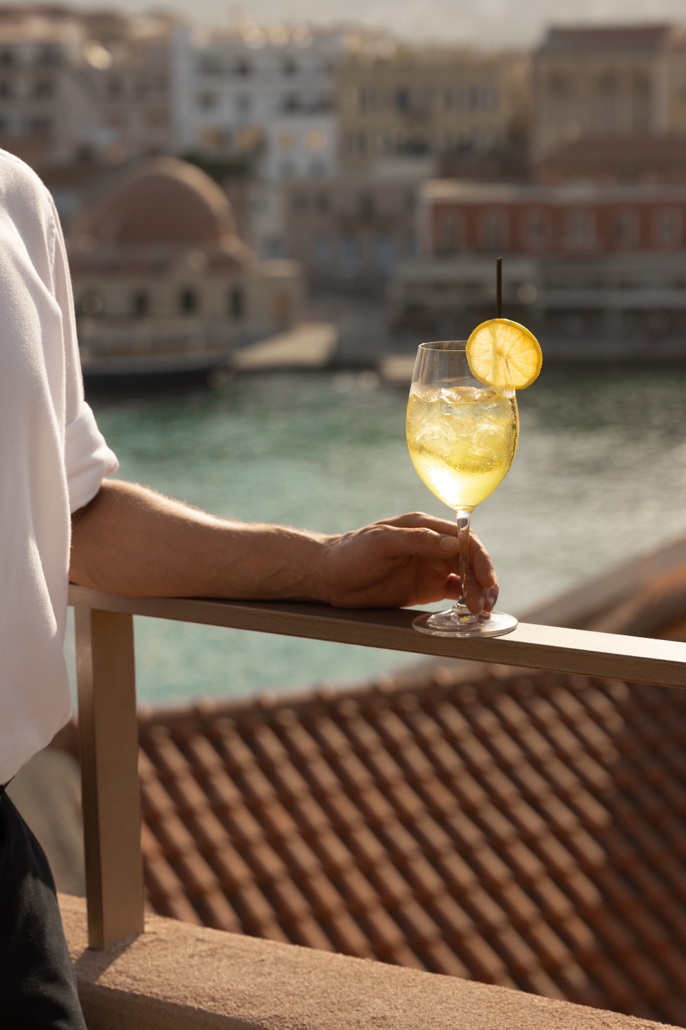 Tall citrus cocktail with an orange wheel garnish resting on a rail above the Venetian harbour, Casa Delfino rooftop bar, Chania, Crete, drinks photography
