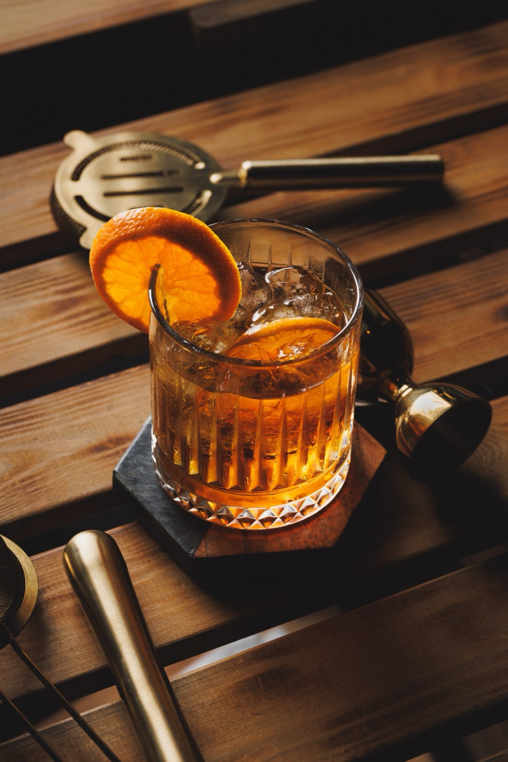 Old fashioned in a cut-crystal tumbler with a curl of orange peel over the rim, brass jigger and bar tools on slatted wood, Padharo, Southampton, cocktail photography