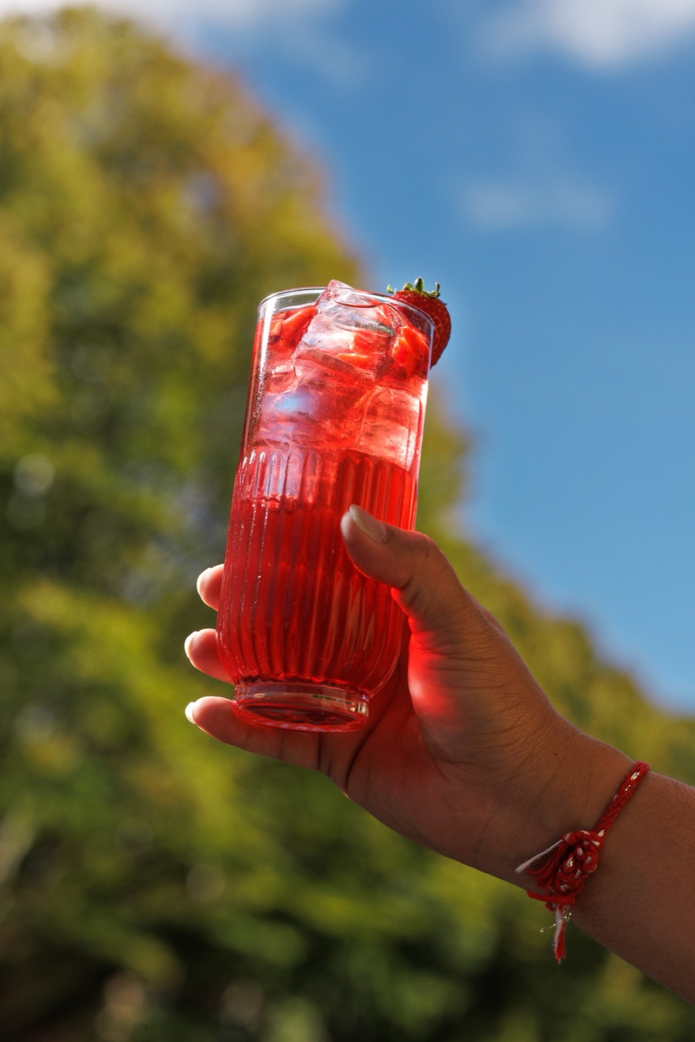 Hand raising a tall strawberry cocktail packed with ice against a blue sky and trees, Padharo, Southampton, cocktail photography