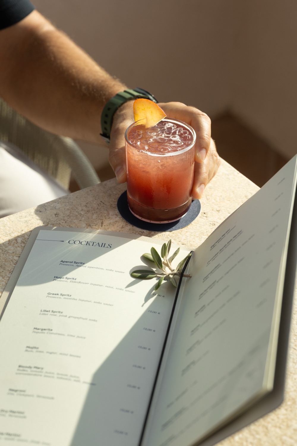 Hand setting down a red cocktail with an orange garnish beside an open cocktail menu on a sunlit stone table, Casa Delfino rooftop bar, Chania, Crete, drinks photography