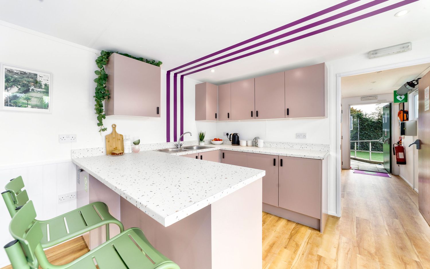 Dining room with pink walls, green marble-topped kitchen island, oak dining table and cane-back chairs, colourful woodland artwork, patterned curtains and slatted pendant light, interior styling photography