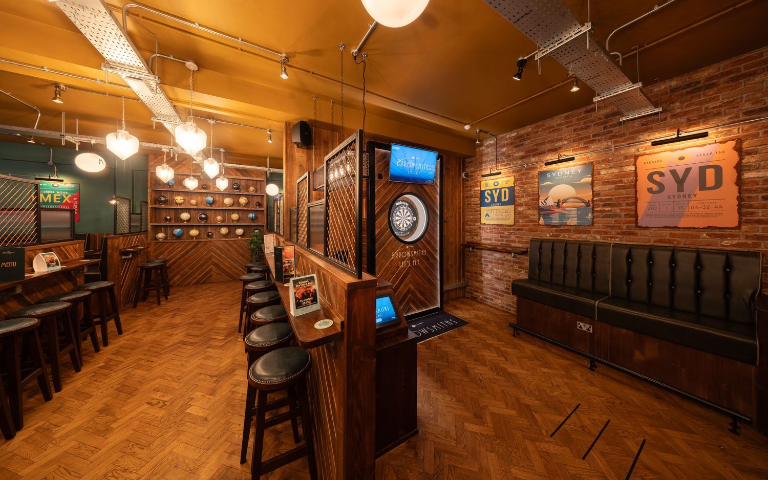Arrowsmiths bar interior with exposed brick walls, parquet flooring, wooden counter with bar stools, electronic dartboard, travel posters and warm pendant lighting, commercial bar photography