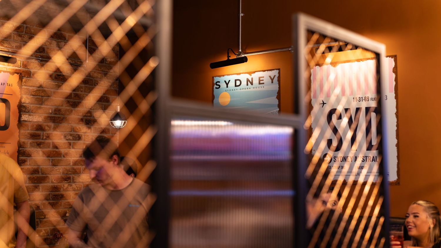 Arrowsmiths bar entrance detail through lattice screen partition with travel posters, exposed brick wall and warm lighting, commercial hospitality interior photography